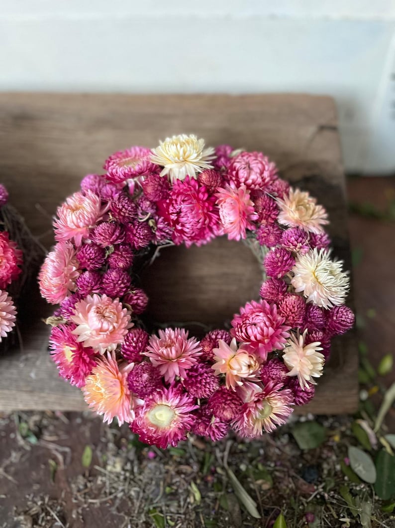 May include: A circular floral wreath composed of various shades of pink and cream-colored dried flowers. The wreath is arranged in a dense, full circle, with a mix of textures and sizes. The flowers are set against a natural, neutral background.