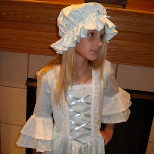 May include: A young girl wearing a white dress with a blue lace bodice and a white bonnet with blue trim. The dress has a ruffled collar and sleeves.