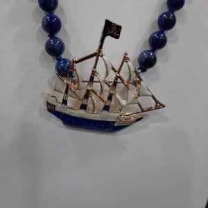 May include: A blue and white ship pendant with a gold trim hangs from a blue lapis lazuli beaded necklace. The ship has a white sail and a black skull and crossbones flag.