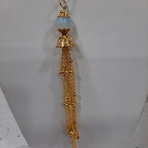 May include: A gold chain necklace with a white opal pendant and a gold tassel. The tassel is made of multiple strands of gold chain with small gold beads.