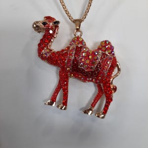 May include: A red camel pendant necklace with a gold chain. The camel is adorned with red and iridescent rhinestones. The pendant has gold-colored feet and a gold-colored head.