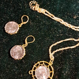 May include: A gold-toned jewelry set consisting of a necklace and two earrings. Each piece features a silver-colored coin with a profile portrait. The necklace pendant is encased in a circular gold frame with decorative spokes, while the earrings have lever-back closures.