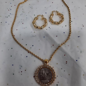 May include: A gold rope chain necklace with a pendant featuring a silver coin with a woman's face and a crown. The pendant is surrounded by clear rhinestones. The set also includes a pair of gold hoop earrings.