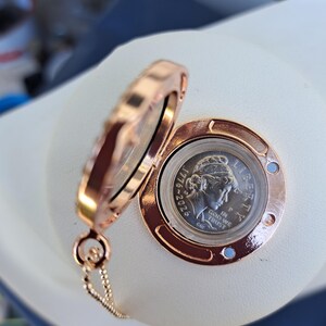 May include: A rose gold-toned circular locket pendant with a hinged opening, containing a silver-colored coin featuring a profile portrait, the text LIBERTY, IN GOD WE TRUST, and the dates 1776-2026, attached to a delicate matching metal chain.
