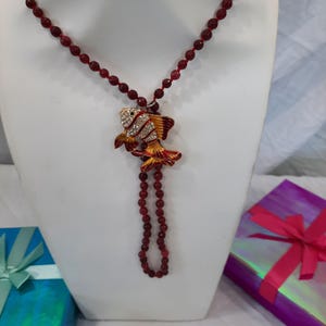 May include: A red beaded necklace with a gold and red fish pendant. The pendant has white rhinestones on it.