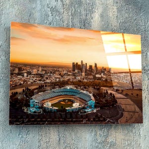 以下が含まれることがあります： ロサンゼルス、カリフォルニア州の野球場の夕暮れの航空写真。スタジアムは、街のスカイラインと大きな駐車場に囲まれています。空は鮮やかなオレンジとピンクです。