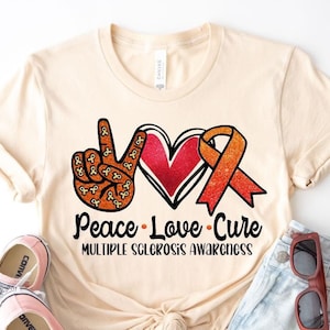 Puede incluir: Una camiseta beige con un signo de paz, un coraz&oacute;n y una cinta naranja con el texto "Peace Love Cure" y "Multiple Sclerosis Awareness".