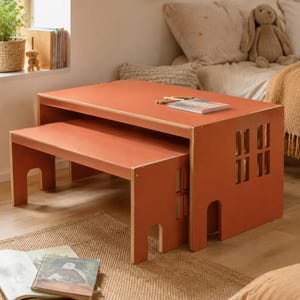 Può includere: Un set tavolo e panca in legno color terracotta con sagome di finestre e porte ritagliate sui pannelli laterali. Il tavolo ha un piano rettangolare piatto e la panca più piccola si inserisce sotto, entrambi in compensato liscio verniciato.