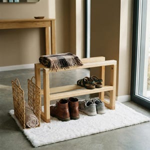 Puede incluir: Un zapatero de madera clara con dos estantes, que contiene varios zapatos y una manta doblada. Dos cestas tejidas y una alfombra blanca están frente al zapatero. El zapatero está cerca de una ventana.