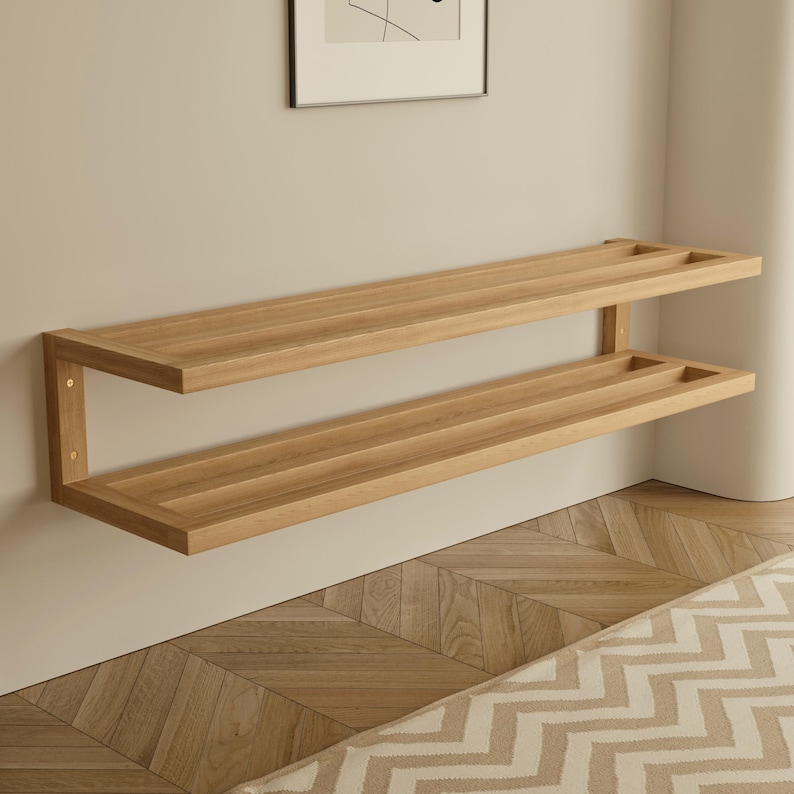 May include: A two-tiered, light-colored wooden wall shelf. The shelves are rectangular with a recessed area on each end. The shelf is mounted on a light-colored wall. The wood grain is visible. The shelf is a modern design.
