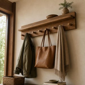Könnte beinhalten: Ein Wandgarderobe aus Holz mit einem Regal, mit sechs Haken zum Aufhängen von Gegenständen. Eine braune Ledertasche, eine grüne Jacke und ein beiger Schal hängen an den Haken. Eine kleine Vase mit Grünzeug steht auf dem Regal.