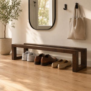 May include: A dark brown wooden bench with a rectangular frame, holding several pairs of shoes. The shoes include white sneakers, brown leather dress shoes, and beige loafers. A canvas tote bag hangs on the wall.