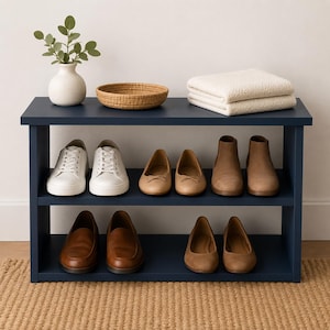 May include: A dark blue two-tier shoe rack holding white sneakers, tan ballet flats, brown ankle boots, brown loafers, and beige flats. The top shelf features a white ceramic vase with green leaves, a woven bowl, and a folded white blanket.