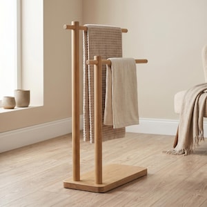 Puede incluir: Toallero de madera con dos toallas beige. El soporte es de madera clara y tiene una base rectangular. Las toallas son de diferentes texturas y tonos beige. El soporte está en una habitación con una ventana y una silla.