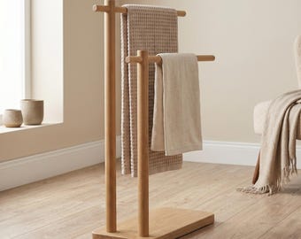 May include: Wooden towel stand with two beige towels. The stand is made of light-coloured wood and has a rectangular base. The towels are different textures and shades of beige. The stand is in a room with a window and a chair.