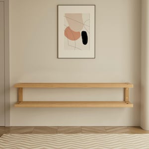 May include: A light-colored wooden wall shelf with two levels is mounted on a beige wall. Above the shelf is a framed abstract art print with pink, black, and beige shapes. The room has a neutral color palette.