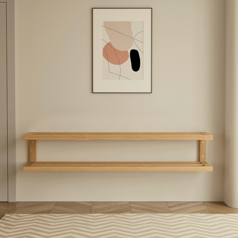 May include: A light-colored wooden wall shelf with two levels is mounted on a beige wall. Above the shelf is a framed abstract art print with pink, black, and beige shapes. The room has a neutral color palette.