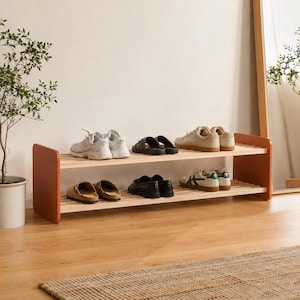 May include: A two-tiered wooden shoe rack with a natural finish and orange-brown side panels. Various pairs of shoes, including sneakers, sandals, and loafers, are displayed on the shelves. The rack is placed on a wooden floor.