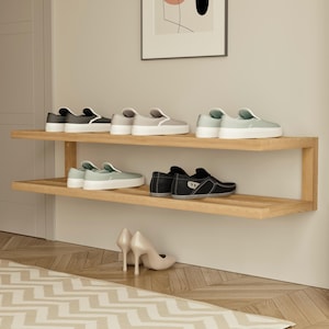 May include: A wooden shoe rack with two shelves displays various pairs of shoes. The shoes include gray, black, and mint green slip-ons, and black leather loafers. A pair of beige high heels sits on the floor below the rack.