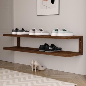 Puede incluir: Un zapatero de madera marrón oscuro de dos niveles montado en una pared blanca. El estante superior contiene zapatillas grises y negras. El estante inferior muestra zapatos de cuero negro. Un par de tacones blancos está en el suelo.