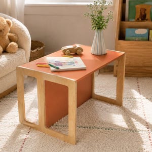 Könnte beinhalten: Ein rechteckiger Kindertisch mit einer glatten, rostorangen Tischplatte und hellen Holzbeinen. Der Tisch hat ein offenes Rahmendesign an den Seiten und trägt ein kleines Holzspielzeugauto, ein Skizzenbuch mit Buntstiften und eine Vase.