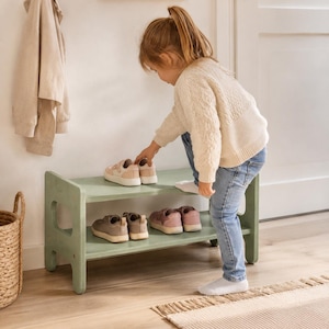 May include: A sage green wooden shoe rack with two horizontal shelves. The rack features a low-profile design with rounded side supports and an open structure, providing storage space for multiple pairs of children's footwear on both levels.