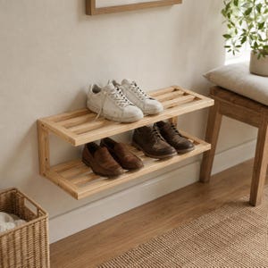 May include: A two-tiered wooden shoe rack mounted on a beige wall. The top shelf holds two pairs of white sneakers, while the bottom shelf displays two pairs of brown leather shoes. The rack is made of light-colored wood.