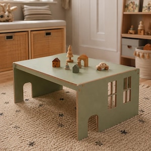 Op de afbeelding: Een saliegroene houten speeltafel ontworpen als een huisje, met boogvormige doorgangen en rechthoekige raamopeningen aan de zijkanten. Op het tafelblad staan diverse kleine houten speelfiguren, waaronder een auto, boompjes en huisjes.
