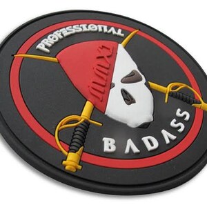 May include: A round, black patch with a red and white design. The patch features a skull-like image with crossed swords and the words "PROFESSIONAL BADASS" in white lettering. The design is set against a black background.