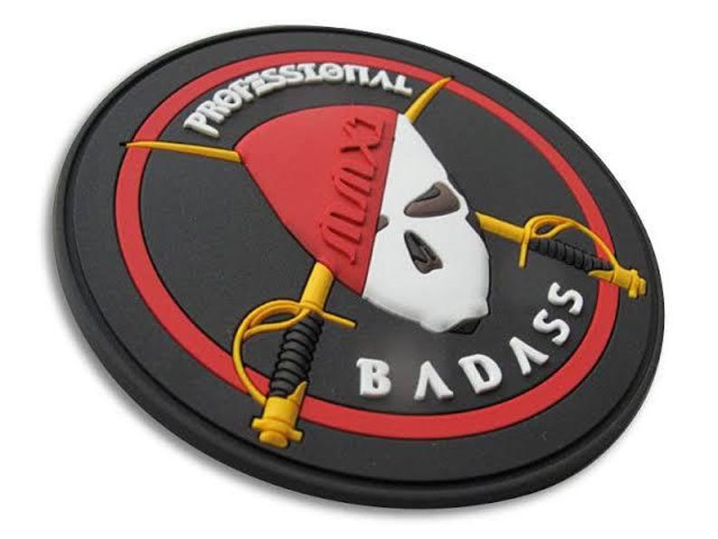 May include: A round, black patch with a red and white design. The patch features a skull-like image with crossed swords and the words "PROFESSIONAL BADASS" in white lettering. The design is set against a black background.