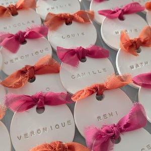 Könnte beinhalten: Weiße Keramik-Namensschilder mit geprägten Namen, verziert mit rosa und orangefarbenen Stoffschleifen. Ideal für Tischkarten, die Etiketten tragen Namen wie VERONIQUE und THOMAS. Anordnung in Reihen.