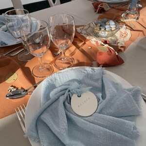 以下が含まれることがあります： 白いお皿に水色のナプキン、ワイングラス、青いキャンディーの小さなボウルが置かれたテーブルセッティング。 テーブルはオレンジと白のリネンで装飾され、小さな赤いポーチが置かれています。