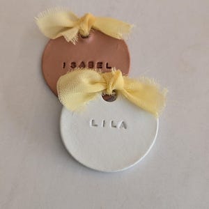 以下が含まれることがあります： 2つの丸いパーソナライズされたネームタグ。1つは「LILA」という名前の白、もう1つは「ISABEL」という名前の銅色です。どちらのタグにも、上部の穴に通された黄色のリボンが付いています。