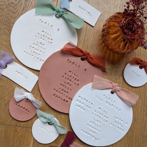 以下が含まれることがあります： 手作りの結婚式用席札のコレクション。白とテラコッタ色の丸型と長方形のカードには、名前とテーブル番号がエンボス加工されています。各カードはカラフルなリボンで飾られています。