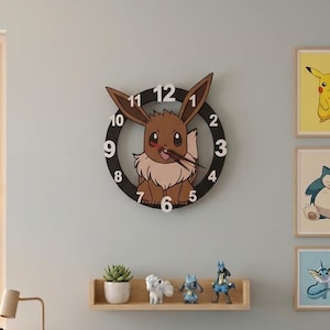 Op de afbeelding: Een ronde wandklok met een bruine Eevee-karakterillustratie met grote oren, witte cijfers van 1 tot 12 en donkere wijzers. De klok heeft een zwart frame en is aan een muur bevestigd boven een houten plank met kleine figuurtjes.