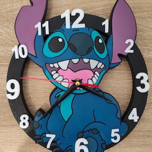 Peut inclure: Une horloge murale circulaire représentant un personnage bleu avec de grandes oreilles et une bouche ouverte montrant des dents. L'horloge possède un cadre noir avec des chiffres blancs de 1 à 12 disposés sur le pourtour. Elle comprend des aiguilles noires pour les heures et les minutes, ainsi qu'une fine trotteuse rouge centrée sur le visage du personnage.