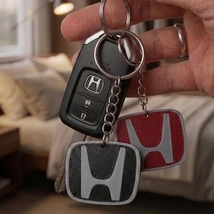 Op de afbeelding: Een zwarte Honda autosleutel met drie knoppen bevestigd aan een metalen sleutelhanger. Twee vierkante sleutelhangers zijn ook aan de ring bevestigd, één met een rood Honda-logo en één met een zwart Honda-logo, beide met zilverkleurige randen.
