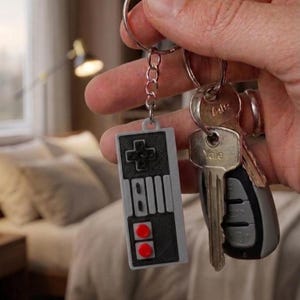 Op de afbeelding: Een rechthoekige grijze en zwarte sleutelhanger in de vorm van een retro videogamecontroller met twee rode ronde knoppen, bevestigd aan een metalen ring samen met een zilveren Yale-sleutel en een grijze en zwarte autosleutel met metalen afwerking.