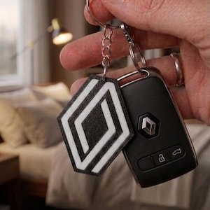 Può includere: Un portachiavi con logo Renault bianco e nero caratterizzato da un emblema a forma di rombo con finitura testurizzata, attaccato a un anello metallico e a una chiave per auto in plastica nera con tre pulsanti e un piccolo logo Renault argentato.