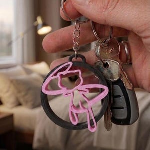 Peut inclure: Un porte-clés circulaire noir présentant le contour rose d'une créature stylisée avec une longue queue et des oreilles pointues, attaché à un anneau métallique avec des clés argentées et un boîtier de clé en plastique gris.