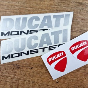 May include: Two white stickers with the text "DUCATI MONSTER" in black and silver lettering. Two red stickers with the text "DUCATI" in white lettering.