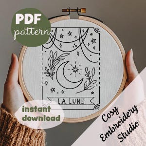 May include: A black and white embroidery pattern featuring a crescent moon, stars, and floral sprigs. The pattern is titled "La Lune" and is displayed in a wooden embroidery hoop.