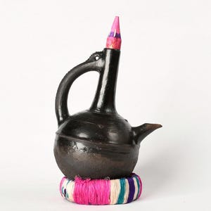 May include: A dark brown, handcrafted ceramic coffee pot with a long spout and handle. The pot has a decorative pink and white striped top and sits on a colorful woven base with pink, white, and blue threads. The pot is likely used for brewing coffee.