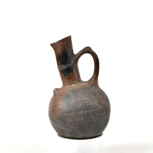 Puede incluir: Una jarra de cerámica marrón oscuro hecha a mano, con un cuerpo redondeado y un pico largo e inclinado. La jarra presenta un asa y líneas grabadas decorativas. El pico y el asa son de un tono marrón más oscuro.