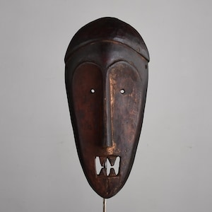 May include: A dark brown, wooden mask with a long, rectangular shape. The mask features a prominent nose, two eye holes, and a mouth with a carved, tooth-like design. The mask is mounted on a metal stand.