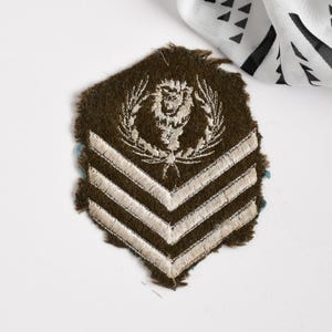 May include: A vintage military patch with a brown background and white embroidered details. The patch features a lion emblem within a laurel wreath and three chevron stripes. The edges are frayed, giving it a worn appearance.