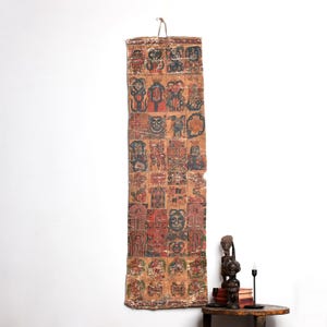 Puede incluir: Textil antiguo rectangular con una cuadrícula de figuras y símbolos estilizados y coloridos. La obra de arte es principalmente en tonos rojos, negros y beige, con un gancho de metal en la parte superior para colgar. Se exhibe contra una pared blanca.