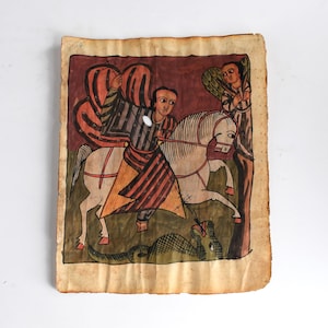 Op de afbeelding: Antieke Ethiopische icoon die een krijger te paard afbeeldt die een slang doodt. Het kunstwerk toont levendige kleuren, waaronder rood, bruin en groen, op verouderd papier. De krijger wordt vergezeld door een engel, tegen een donkere achtergrond.