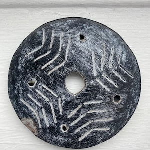 May include: A round, black stone object with a central hole and several smaller holes. The surface is textured with white, linear patterns radiating from the center. The object appears to be weathered, with a rustic aesthetic.