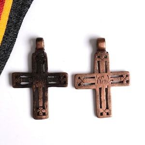 May include: Two metallic cross pendants with a dark bronze finish. Each cross features an openwork design with etched linear patterns and a small loop at the top for hanging. The left cross is darker, while the right cross has a lighter copper tone.